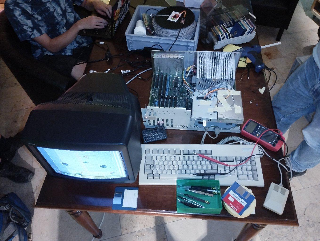 Ein Amiga 2000 mit geöffnetem Gehäuse, und freiem Blick auf die Kabel der Diskettenlaufwerke und das Mainboard. Daneben ein Röhrenfernseher, Tastatur, Maus und Disketten.