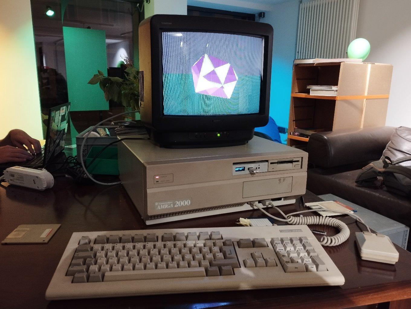 Ein Amiga 2000 mit einem Röhrenfernseher oben drauf, der einen Ausschnitt der “9 Fingers”-Demo zeigt.